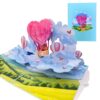 Carte d’amour pop-up 3D ouverte avec une montgolfière en forme de cœur au-dessus des nuages, à côté de sa couverture bleue