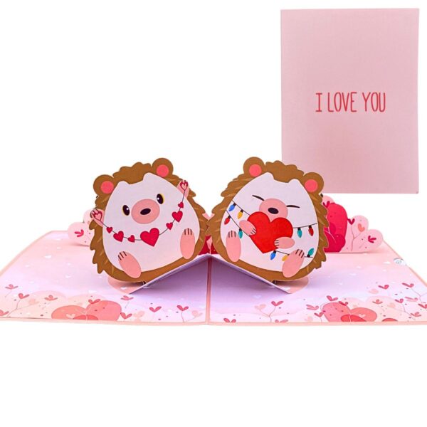 Open 3D pop-up love card with two hedgehogs, a heart garland and fairy lights, next to its pink front cover